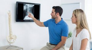 A male chiropractor showing a female patient her neck X-rays on a digital screen, explaining a potential injury from a car accident.